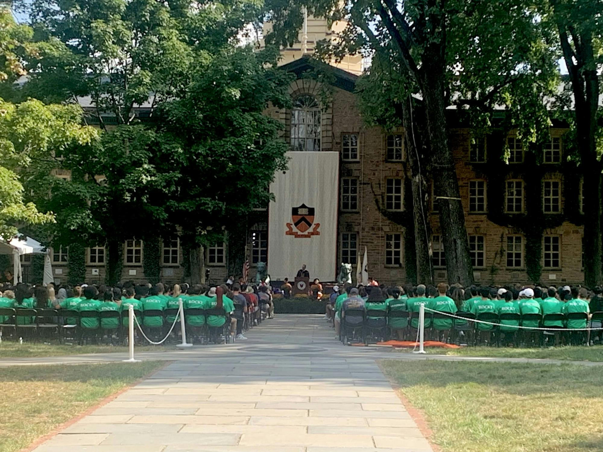 Students in green t-shirts sit in rows of folding chairs in front of an ivy-covered building.