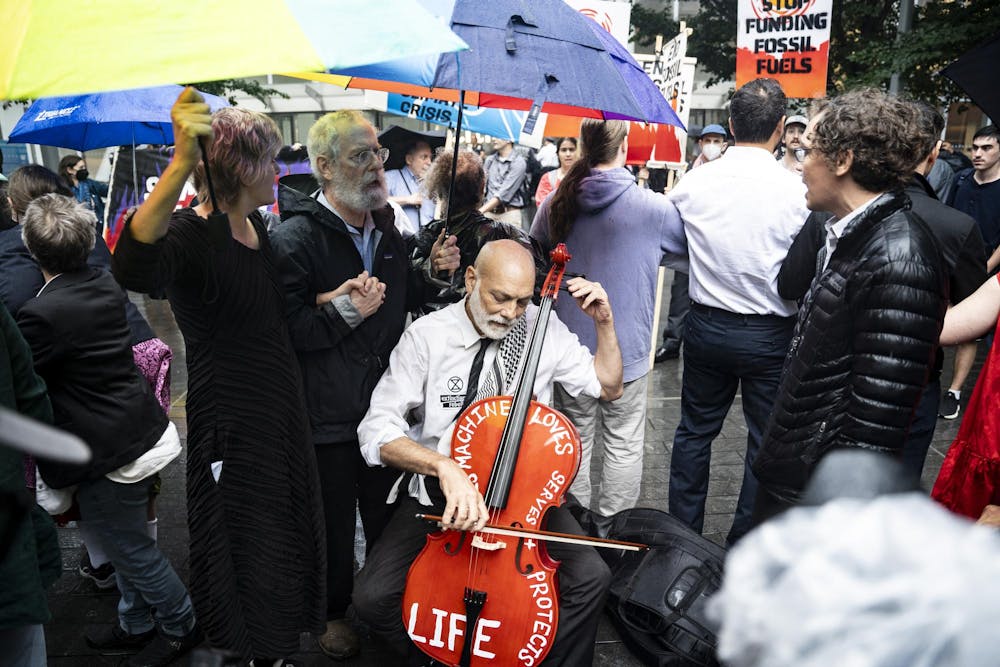 An older man plays the cello in a concrete plaza. On the cello is written, "this machine loves, serves, and protects." A crowd of people stand around the cellist, some holding umbrellas over his head and instrument. 