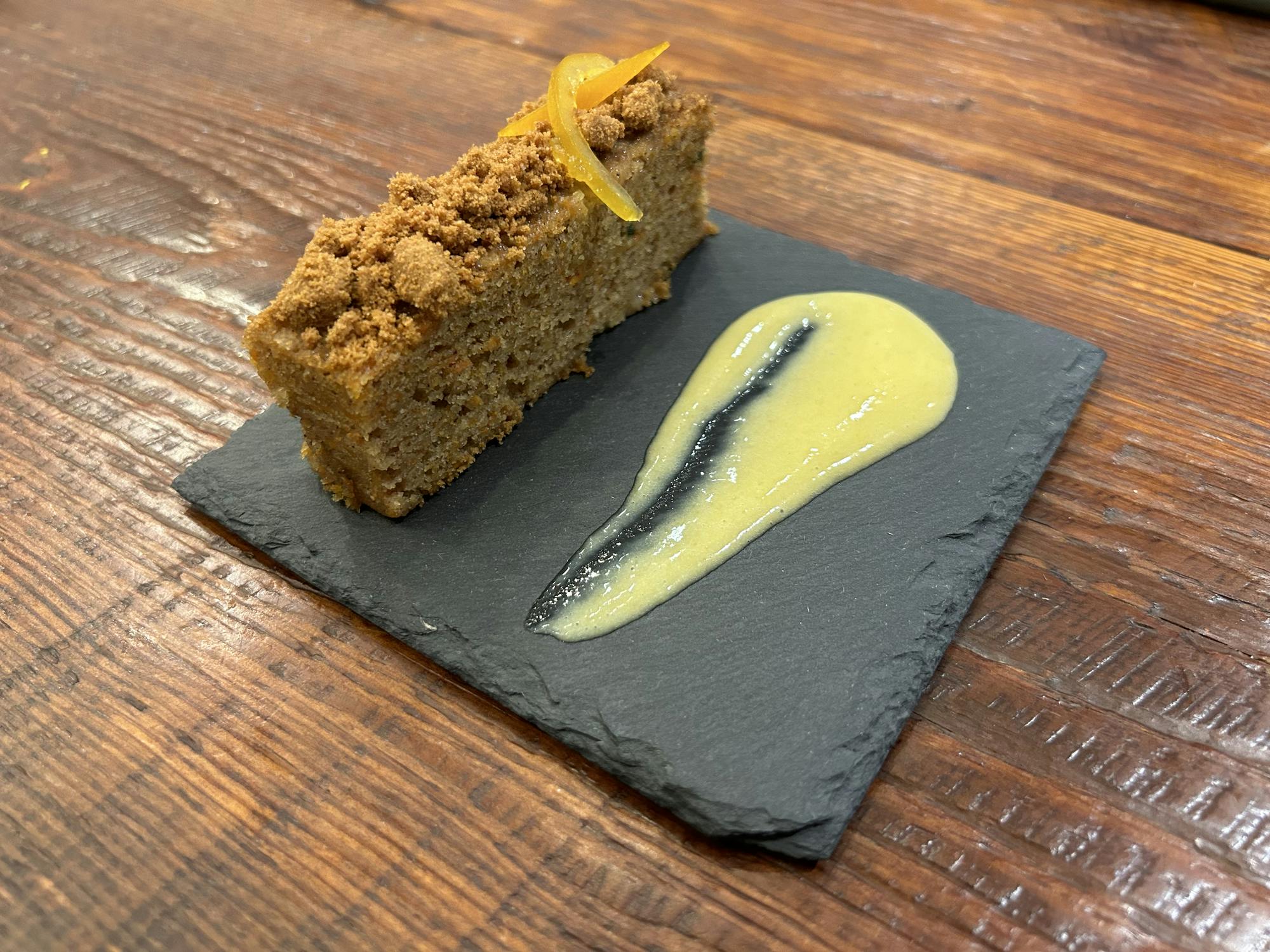 Slice of carrot spice cake, topped with candied orange and biscoff crumble, and a smear of persimmon orange curd on the side.