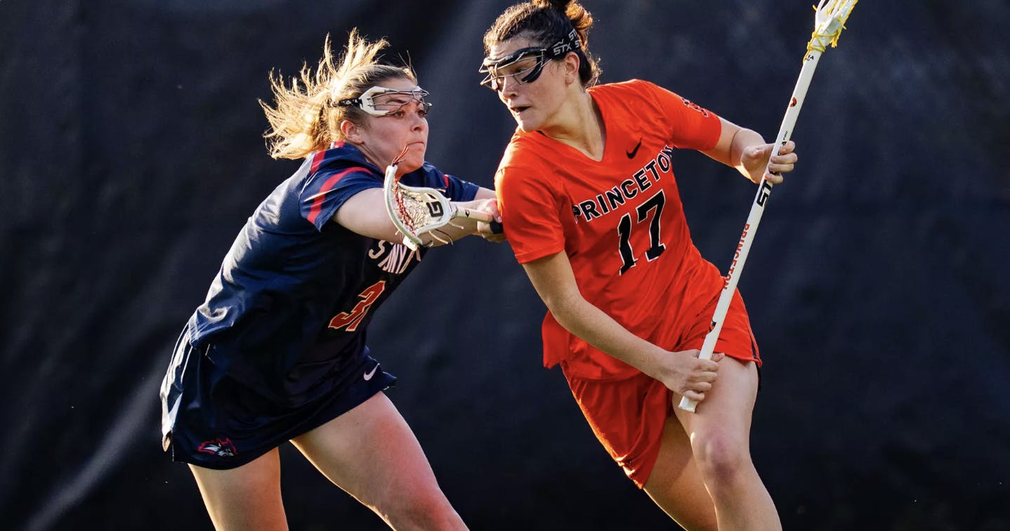 A player in an orange jersey holding a lacrosse stick being defended by a player in a blue jersey holding a lacrosse stick.