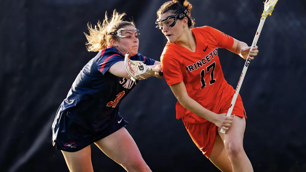 A player in an orange jersey holding a lacrosse stick being defended by a player in a blue jersey holding a lacrosse stick.