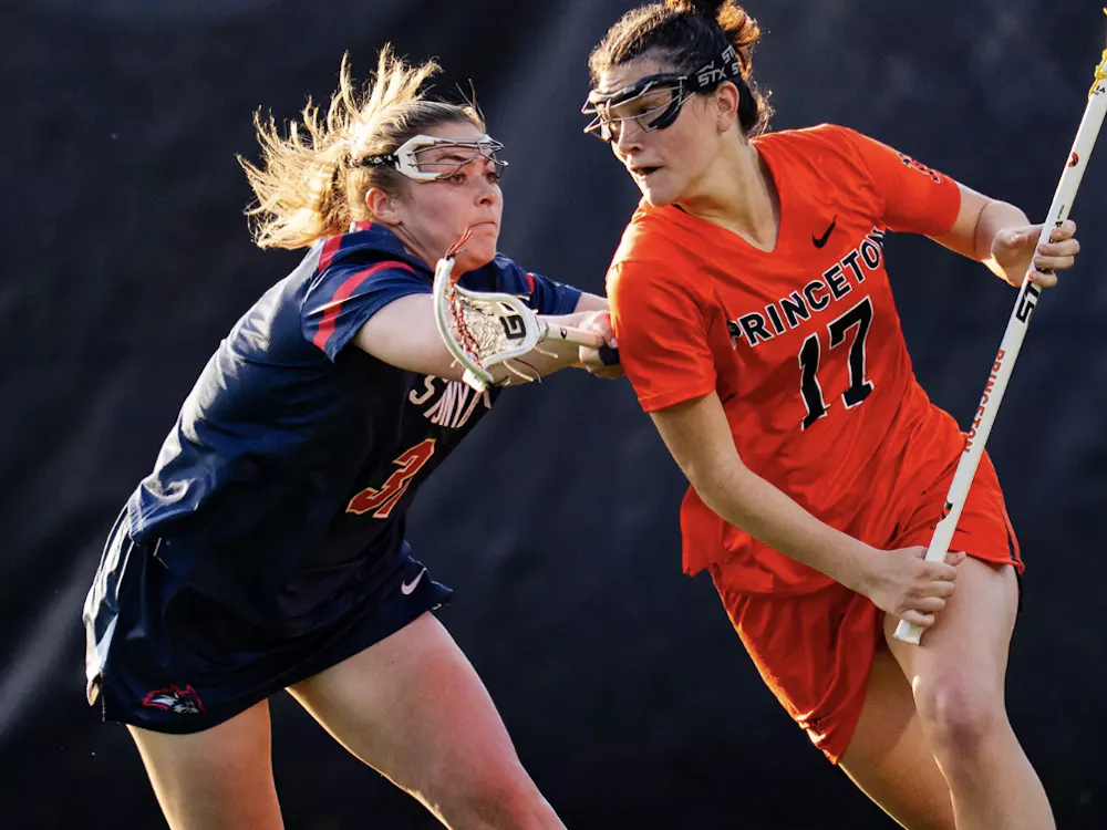A player in an orange jersey holding a lacrosse stick being defended by a player in a blue jersey holding a lacrosse stick.