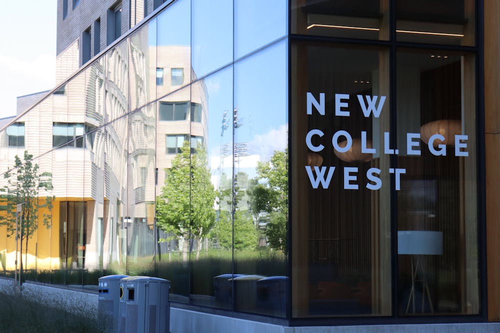 A glass building reflects a blue sky with trees in the background. The building is labeled “New College West.”