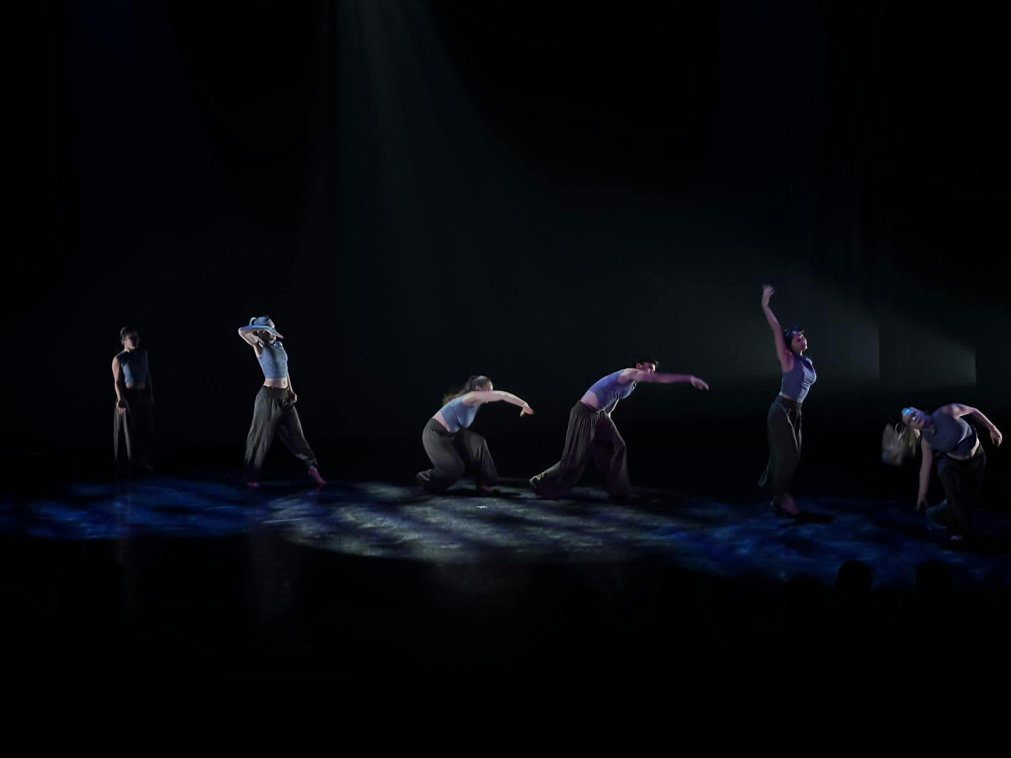 A line of dancers on a black stage form the shape of a wave with their arms.