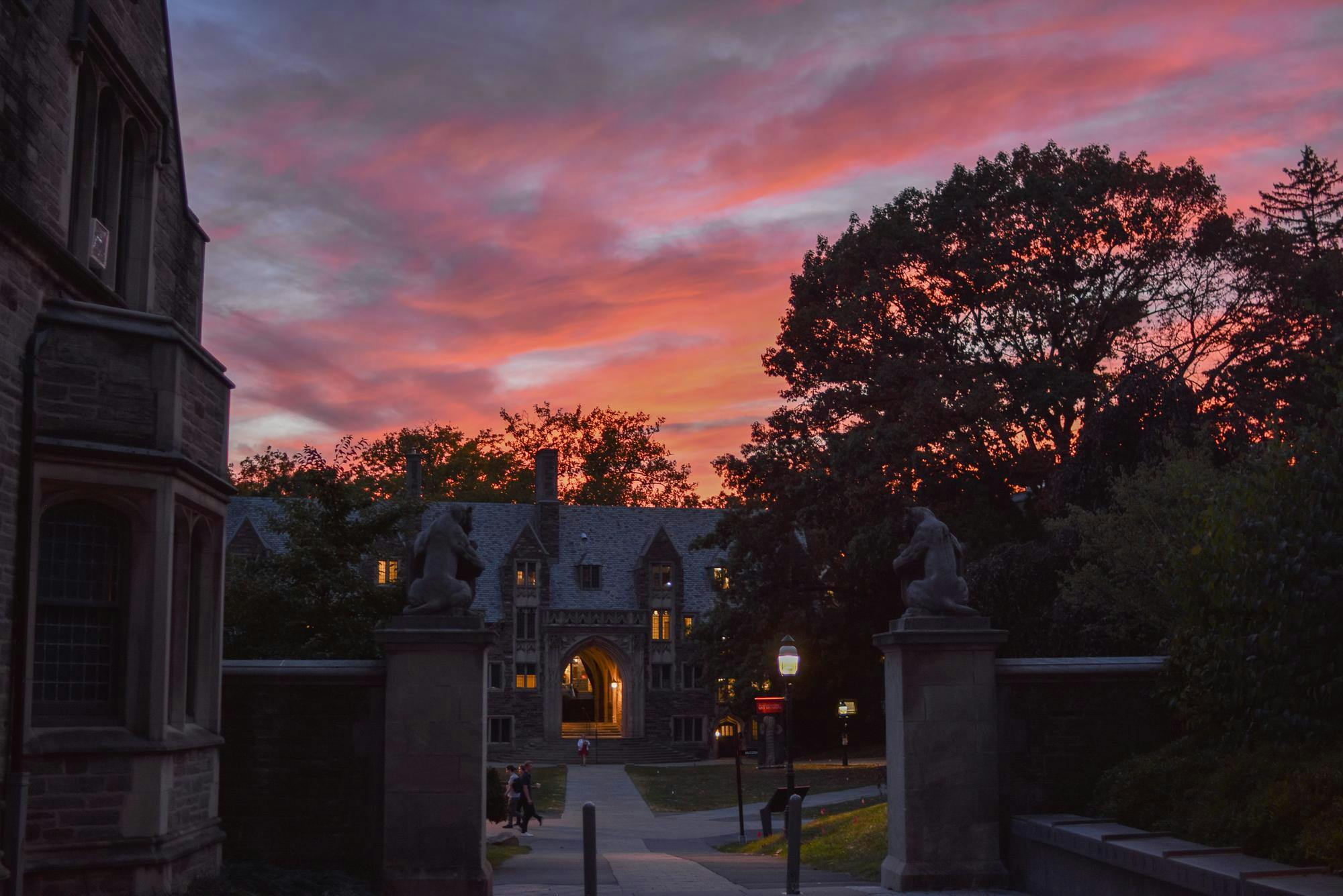 In the foreground, two marble tigers sit perched upon opposite stone grey columns with an open walkway in the middle. In the background, a the stone facade of a dorm building is illuminated in the early dusk. Above, the sky turns from day to night, combining light shades of blue, orange, and purple.