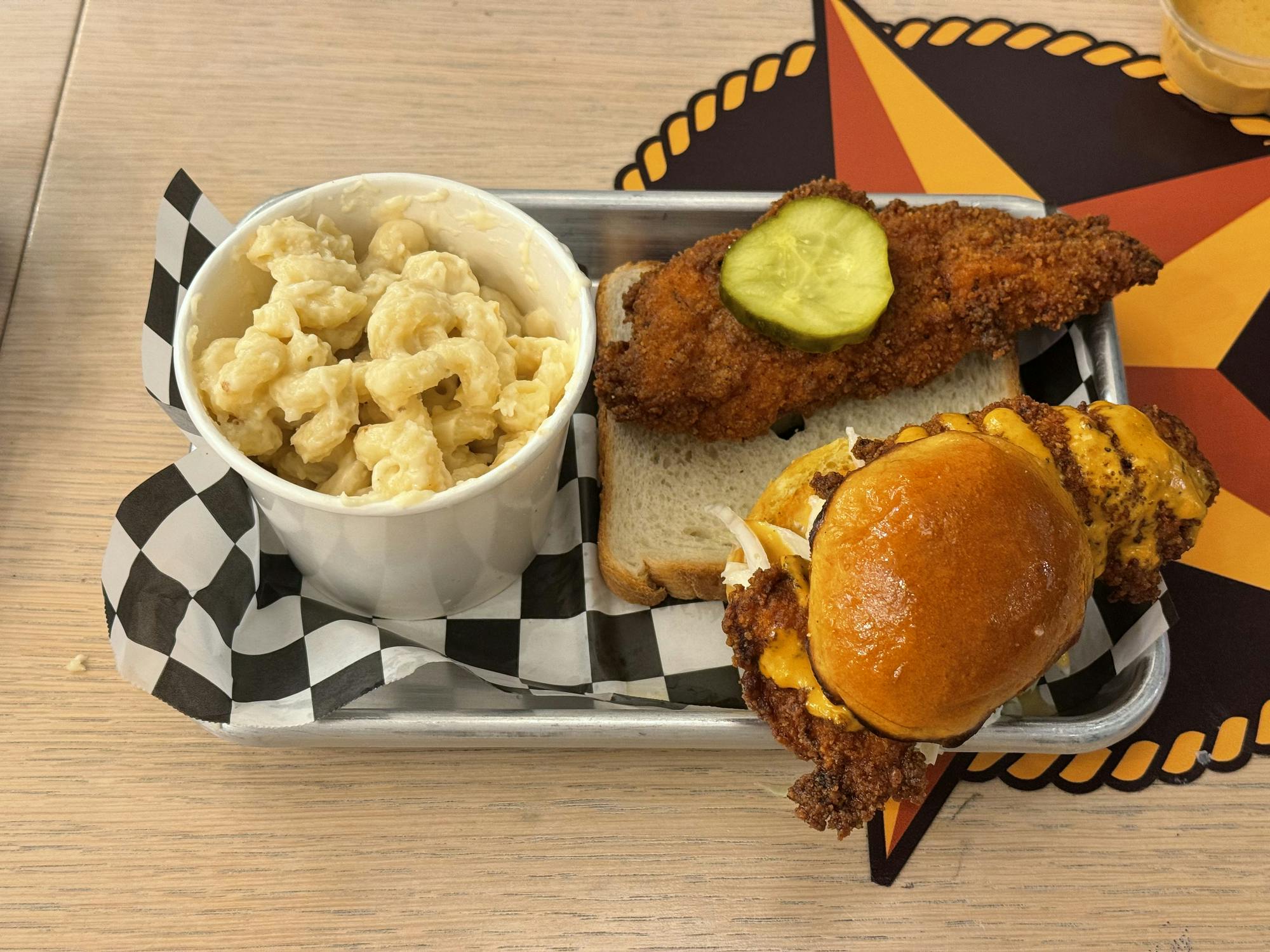 A small grey trey is lined with black and white checkered paper. On top of the tray there is a small white cup with light yellow mac and cheese, a piece of white break with an orange piece of chicken that has a green pickle on top, and a small slider on a toasted bun with orange chicken in the middle of the two halves of the bun.