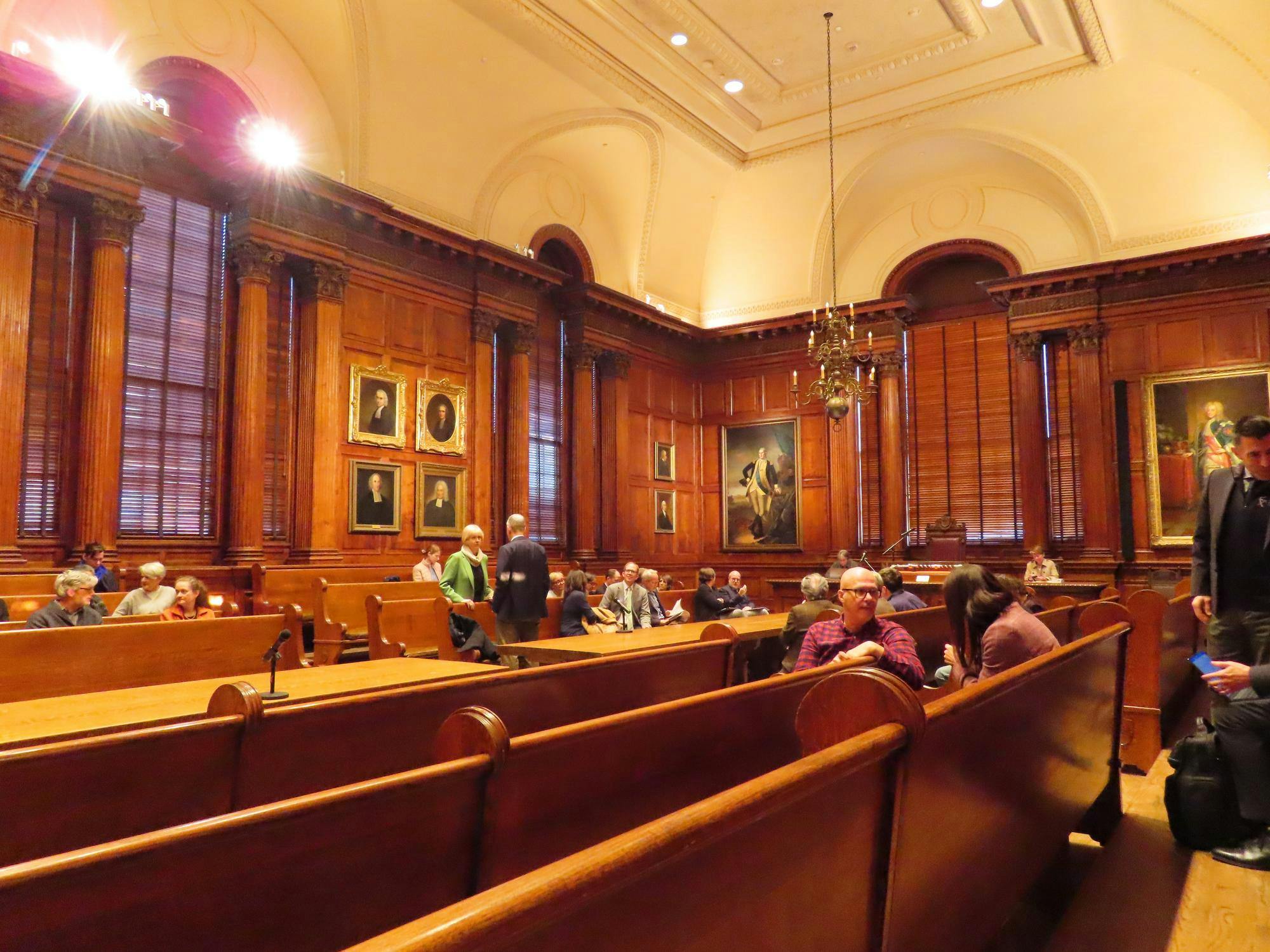 A&#x20;wooden-panelled&#x20;room&#x20;with&#x20;paintings&#x20;on&#x20;the&#x20;walls.&#x20;A&#x20;number&#x20;of&#x20;people&#x20;are&#x20;sitting&#x20;in&#x20;wooden&#x20;benches&#x20;around&#x20;the&#x20;room.