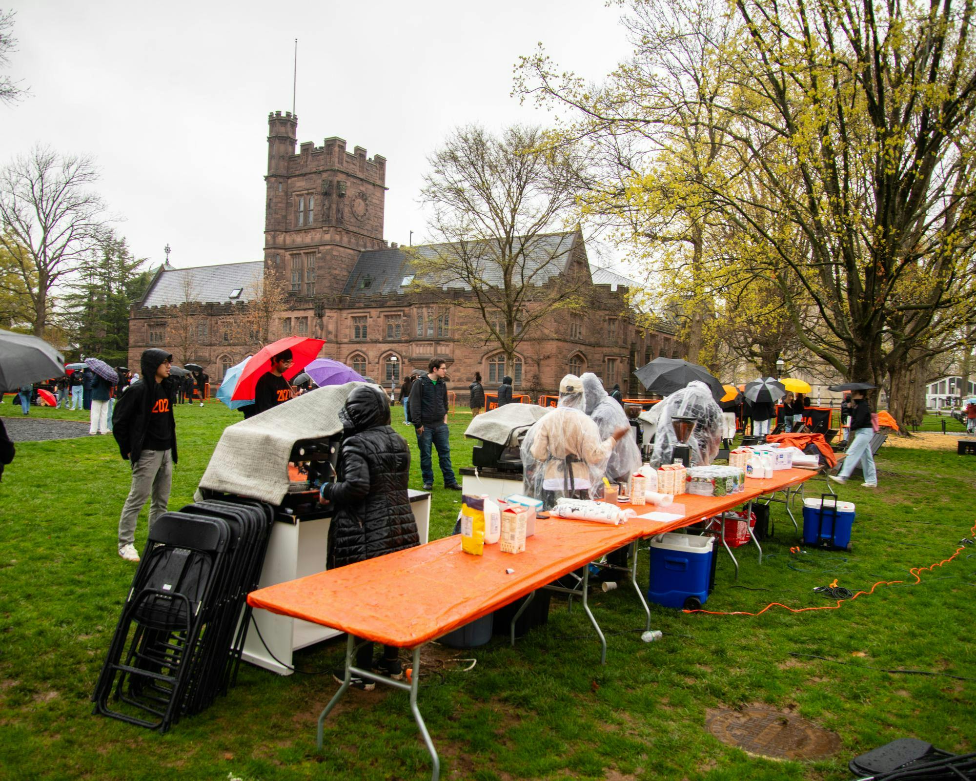 People with umbrellas and ponchos stand behind a table covered with an orange plastic sheet. The table has drinks on it. 