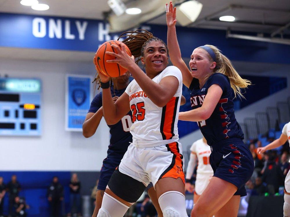 Player in orange and white drives to basket with basketball against two Penn players in blue. 