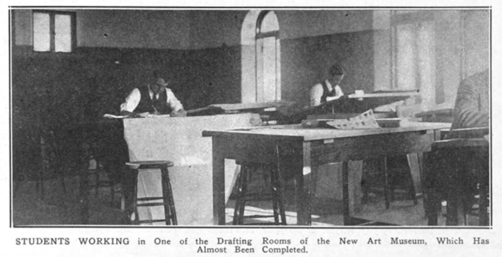 A black and white image of two students in waistcoats and ties, working behind drafting tables illuminated by light from large windows off to the side. 