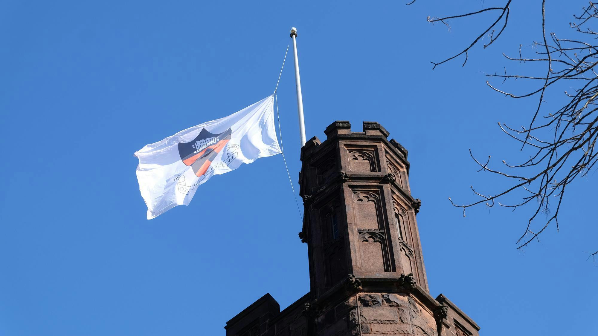 A brown column of a building has a white flag pole on top. A white flag with the orange and black Princeton emblem flies from the pole.