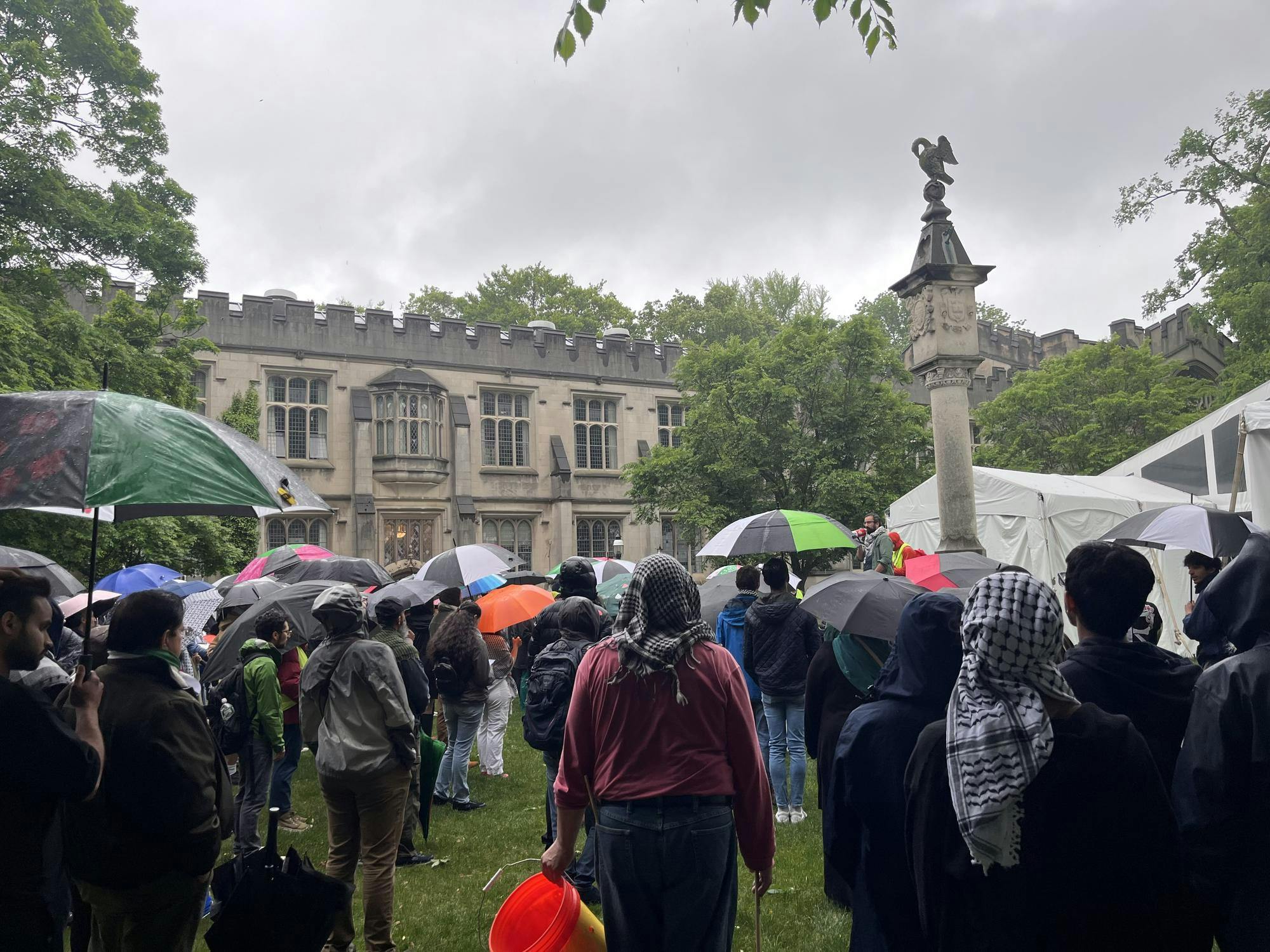 In the front, several individuals face a sundial which a speaker is standing on. In the back, a gray stone building.