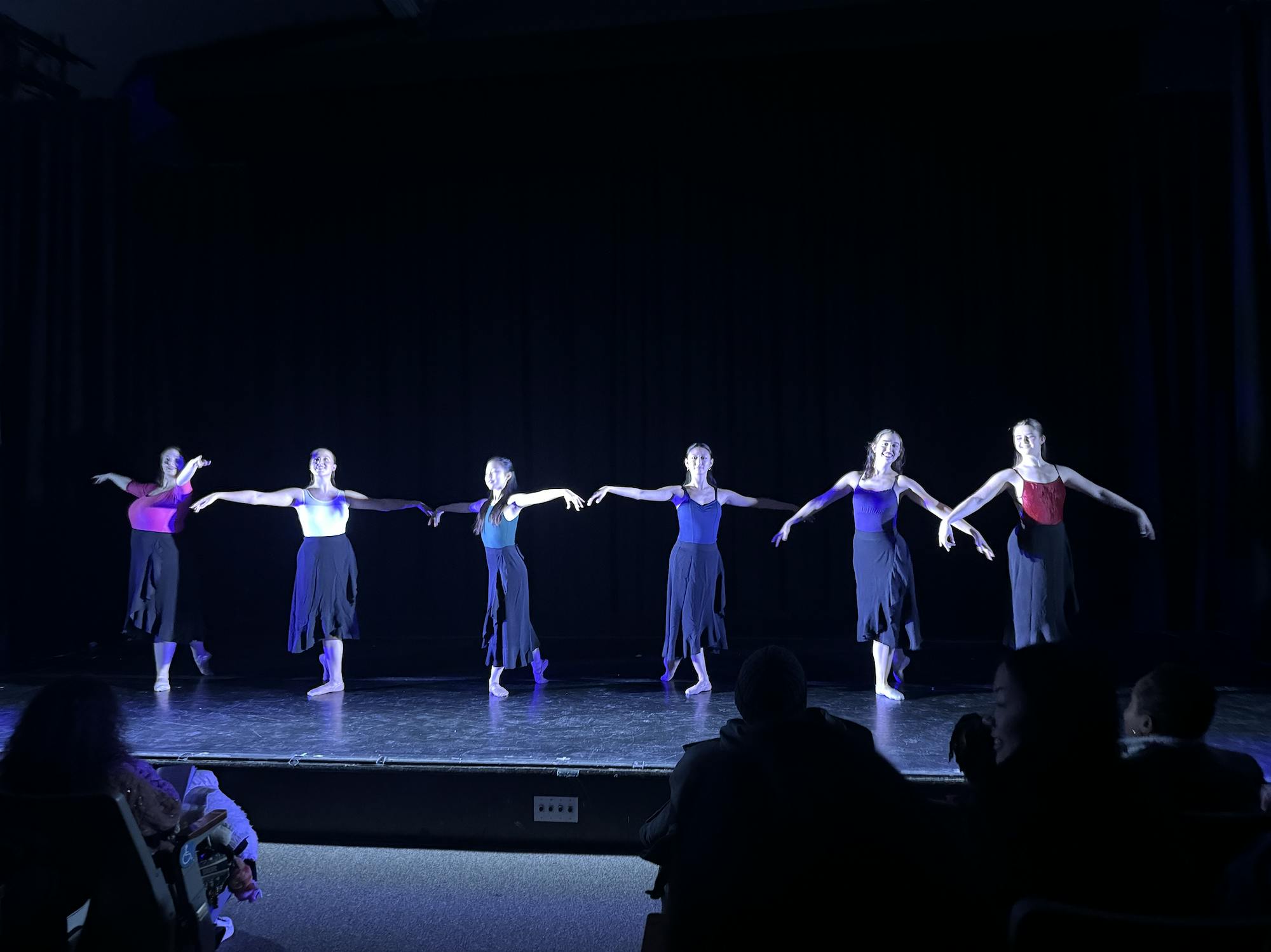 Six ballerinas pose in black clothing and in blue lighting.