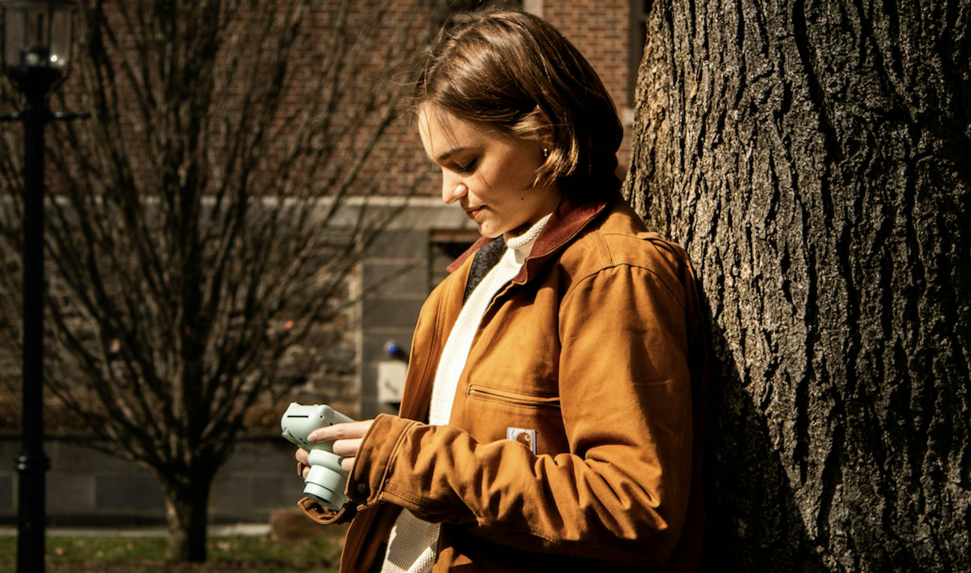 A person wearing a brown jacket with short blonde hair is holding a pastel-colored camera.
