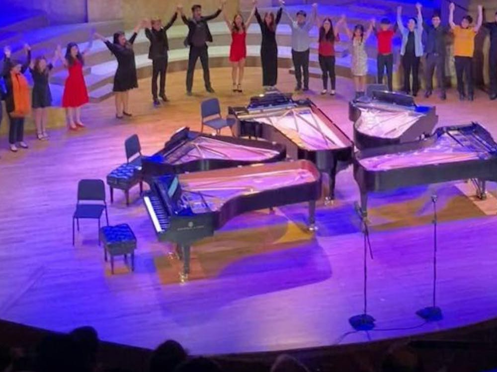 Five grand pianos in the center of a stage. A semi-circle of students stand around the back of the stage with hands held high.