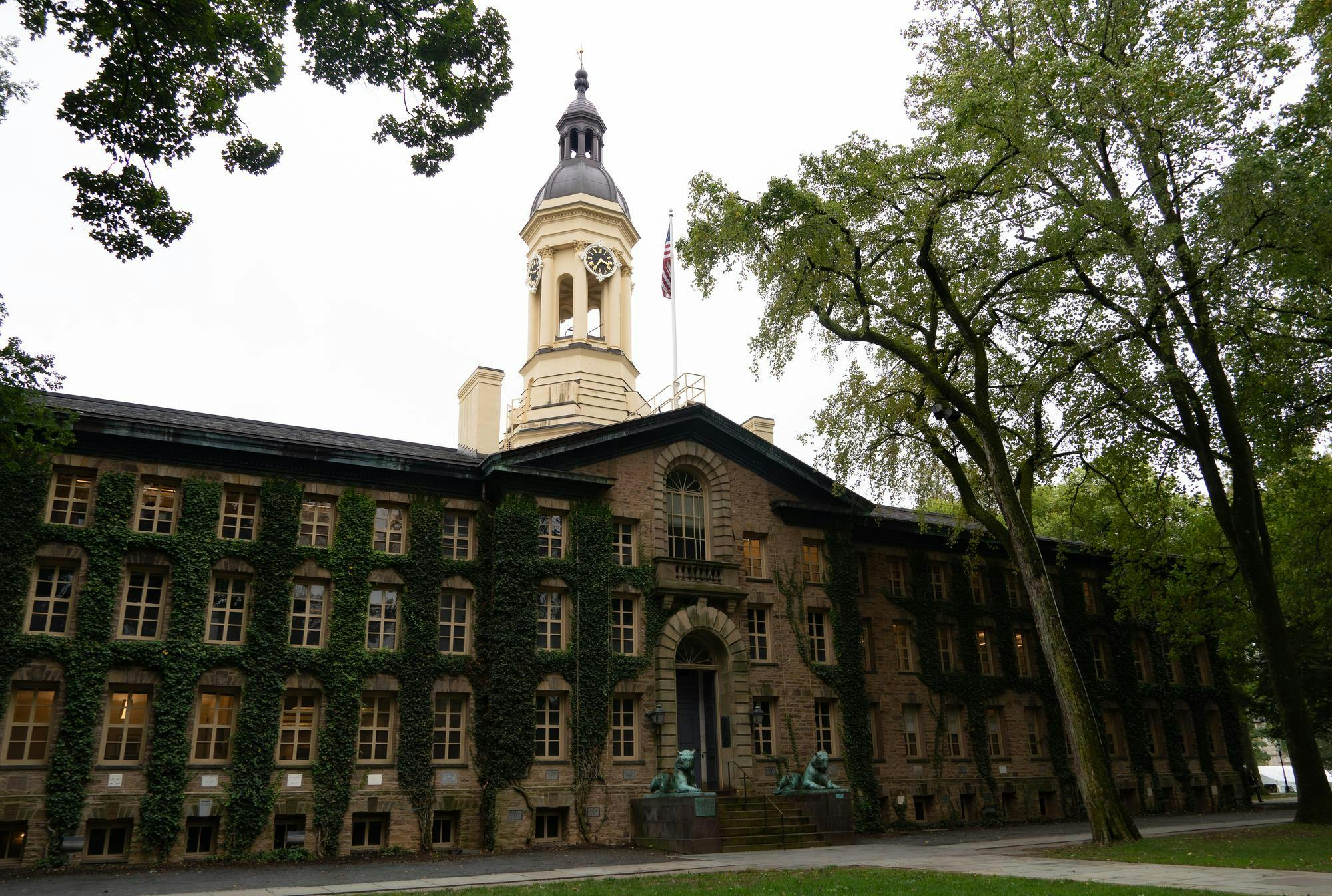 Front profile from the left of Nassau Hall’s front facade. Tan, stone building covered by green ivy, with a clock and bell tower above and an American flag. The building is surrounded by trees, predominately on the left of the image. 