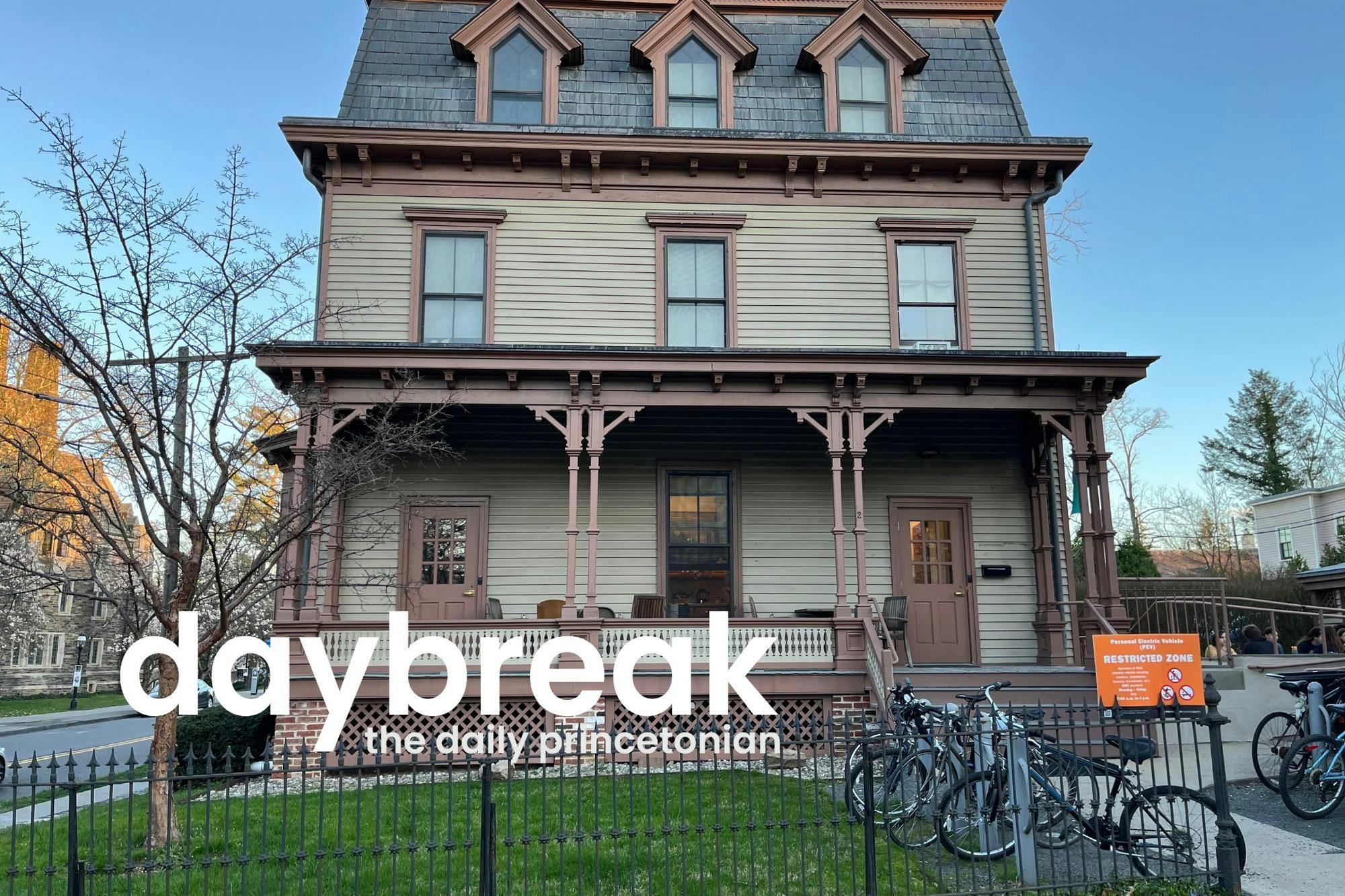 The 2 Dickinson vegetarian co-op at sunset. A red-and-tan 3-floor building with a grey roof. Bikes are parked outside. A logo with text that says "daybreak the daily princetonian" is featured in the bottom left of the image. 