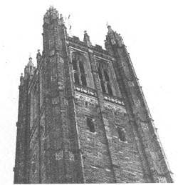 Black and white photograph of the top of Holder Tower, a tall gothic tower made of grey stone that is a part of Rockefeller College.
