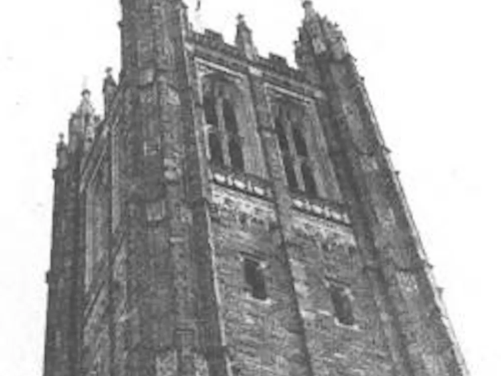 Black and white photograph of the top of Holder Tower, a tall gothic tower made of grey stone that is a part of Rockefeller College.