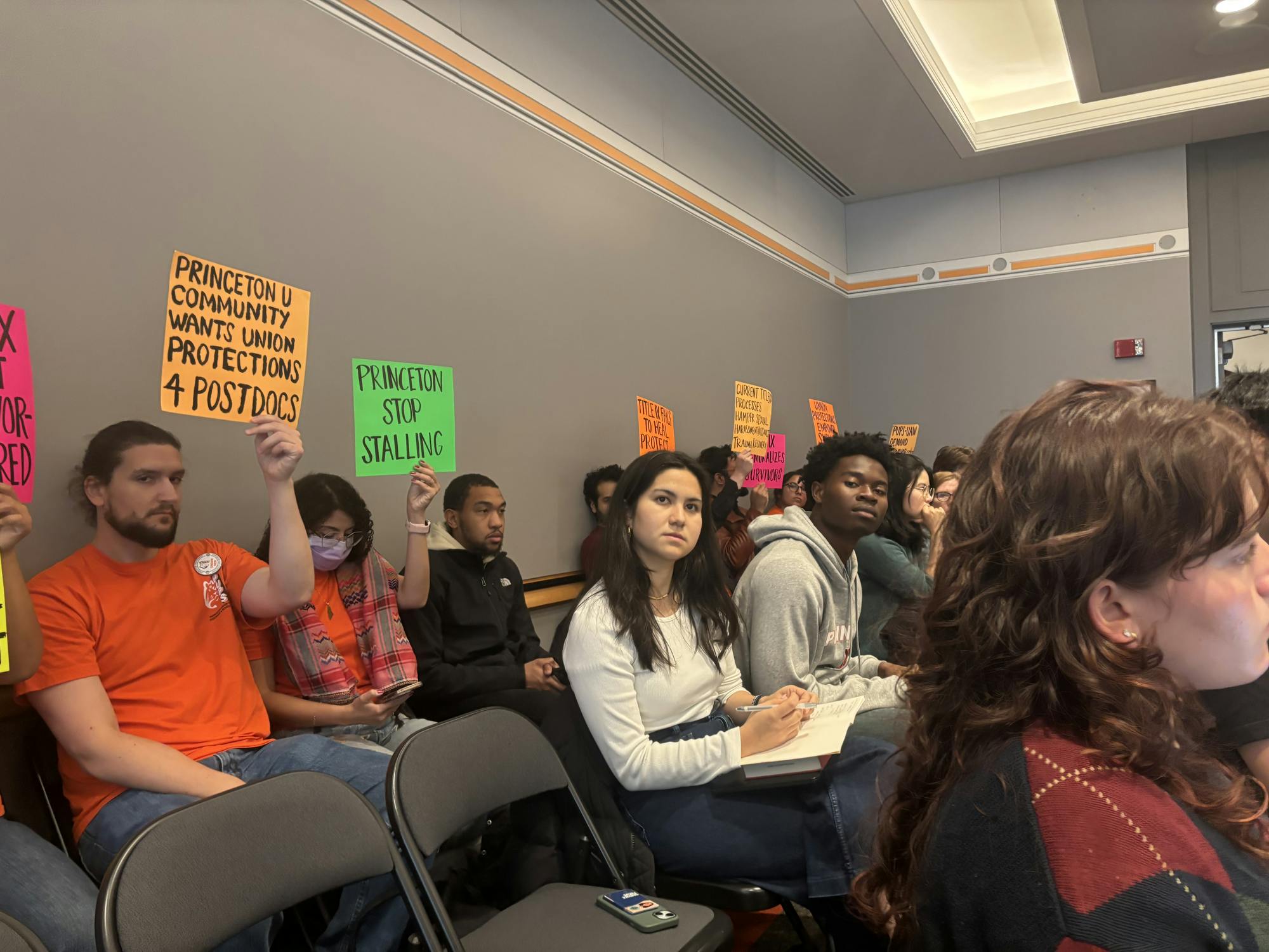 A group of people sitting in an audience in a room with grey walls, holding multicolored signs up.
