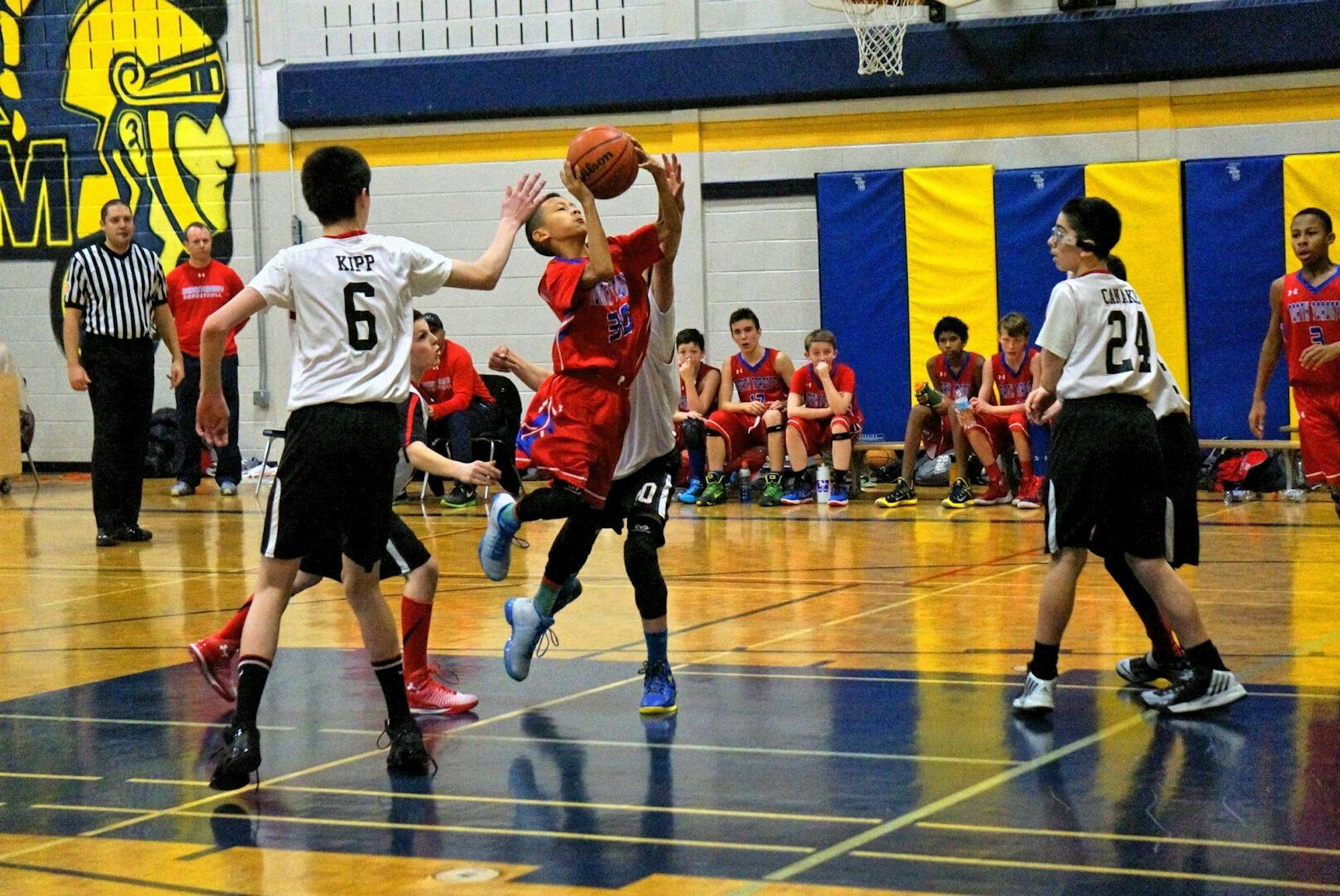 A boy in a red jersey that reads "NORTH TORONTO" and red shorts launches an orange basketball towards a basket. A player in a white jersey, numbered six, tries to block him.