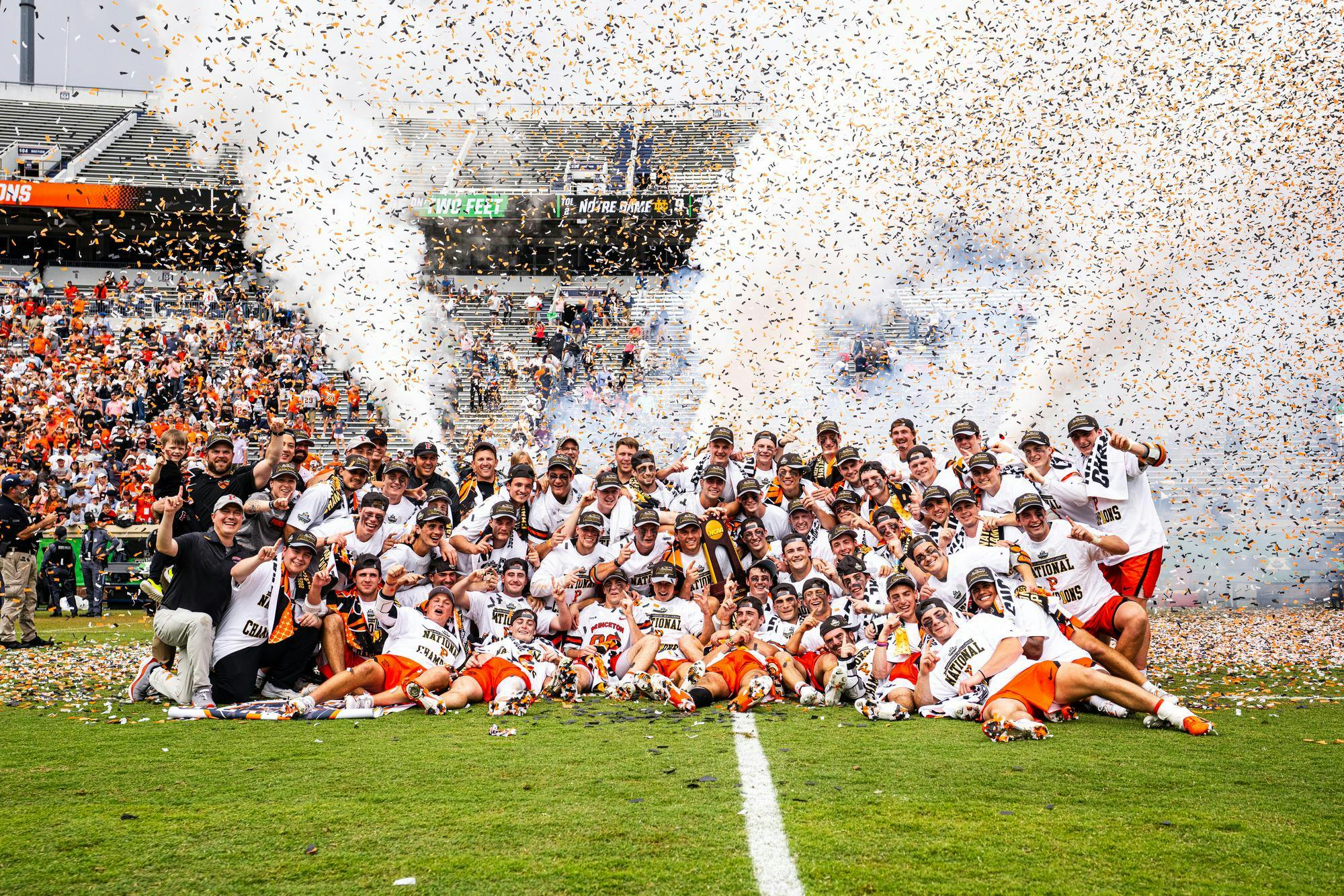 A group of Princeton lacrosse players celebrating on the field with confetti in the background and fans wearing Orange and Black in the stands.
