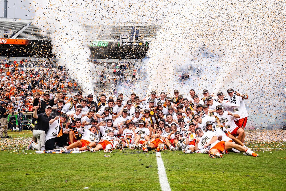 A group of Princeton lacrosse players celebrating on the field with confetti in the background and fans wearing Orange and Black in the stands.
