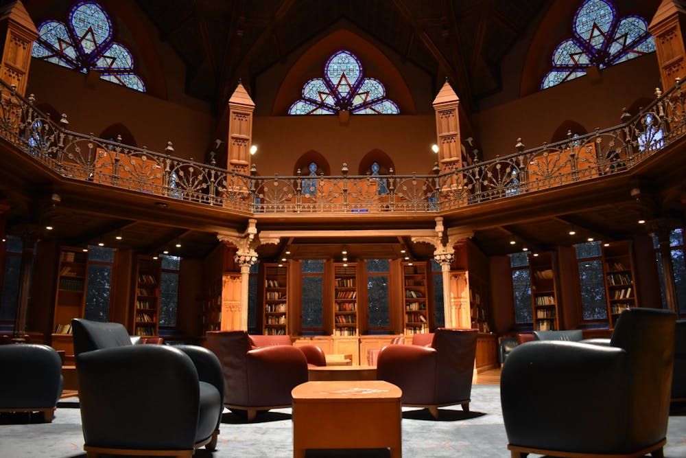 Photo of interior of a library. Shows scattered chairs (blue and red) on a blue rug, with the wooden walls, bookshelves, and stained-glass windows in the background.