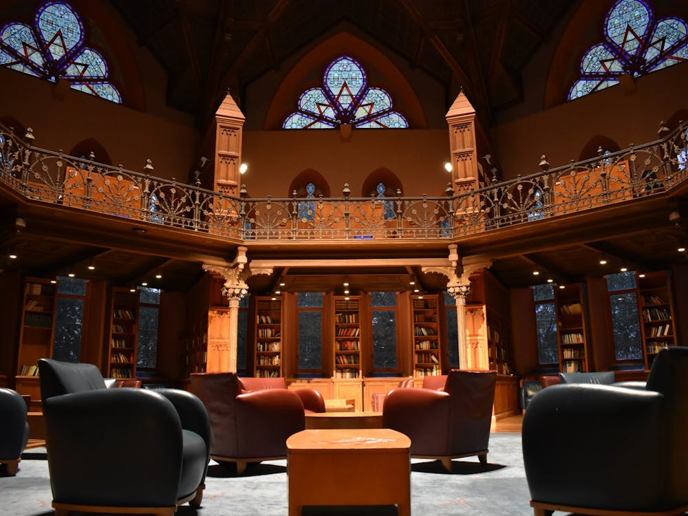 Photo of interior of a library. Shows scattered chairs (blue and red) on a blue rug, with the wooden walls, bookshelves, and stained-glass windows in the background.