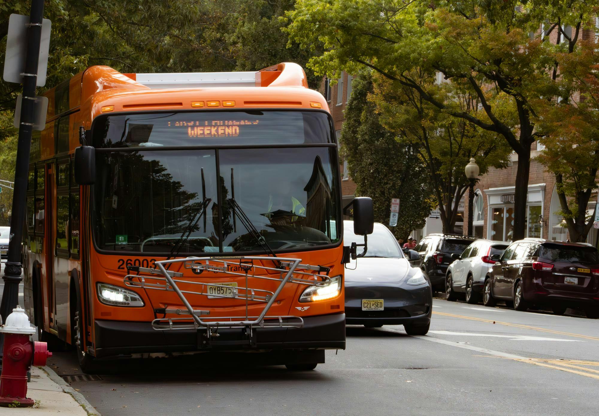 An orange bus shows a sign that reads "First Families Weekend" while parked on the side of a busy road. 
