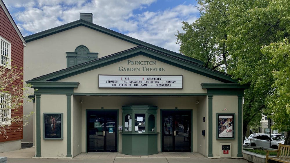 The facade of a movie theater with darker green accents surrounding the doors. A marquee displays coming attractions while the theater itself is flanked by a blue sky and green tree.