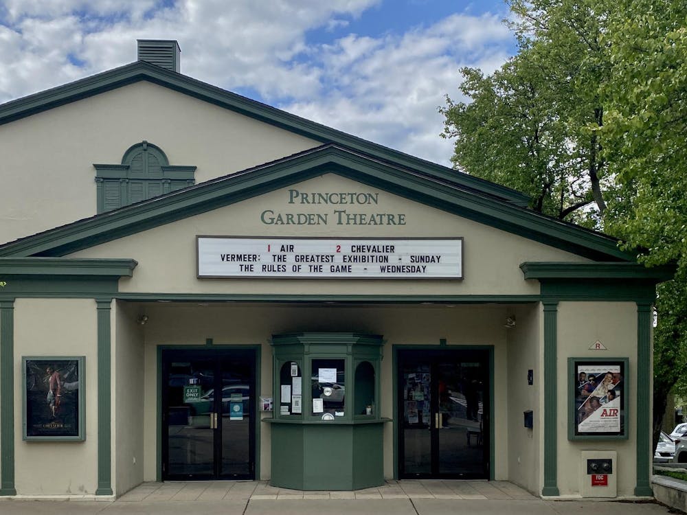 The facade of a movie theater with darker green accents surrounding the doors. A marquee displays coming attractions while the theater itself is flanked by a blue sky and green tree.