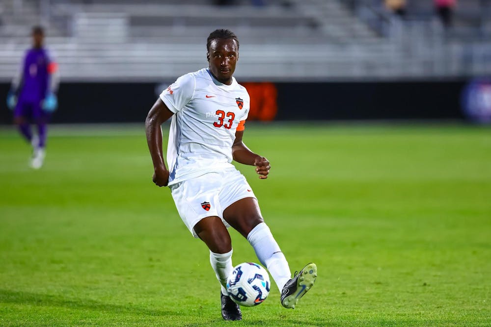 A man wearing white shorts and a white jersey with the number 33, a Nike logo, and a patch all in orange on the jersey on a soccer field kicking a soccer ball. 