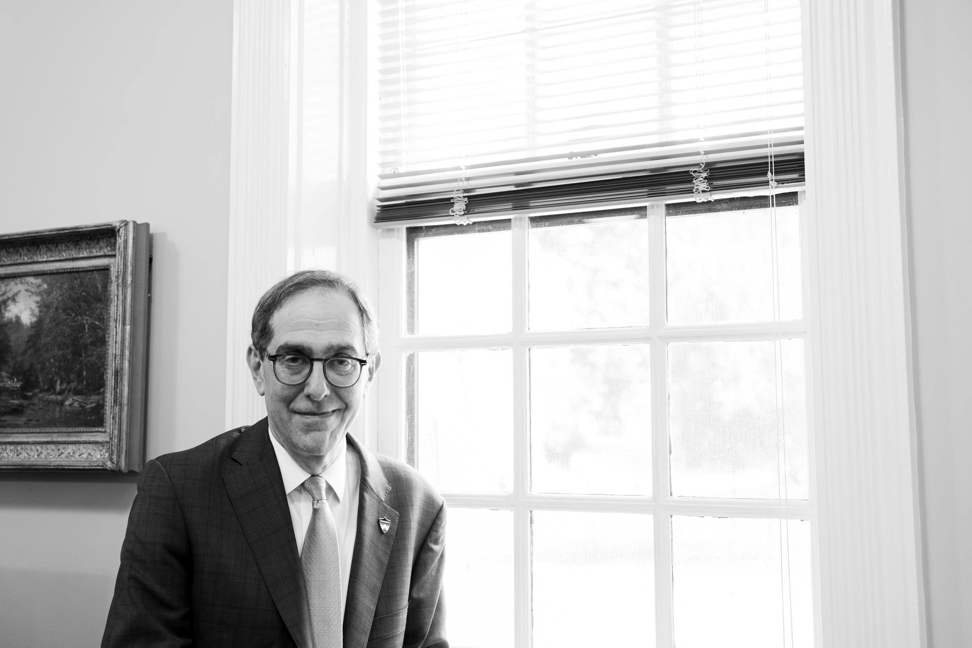 A man with glasses and suit stands in front of a window.