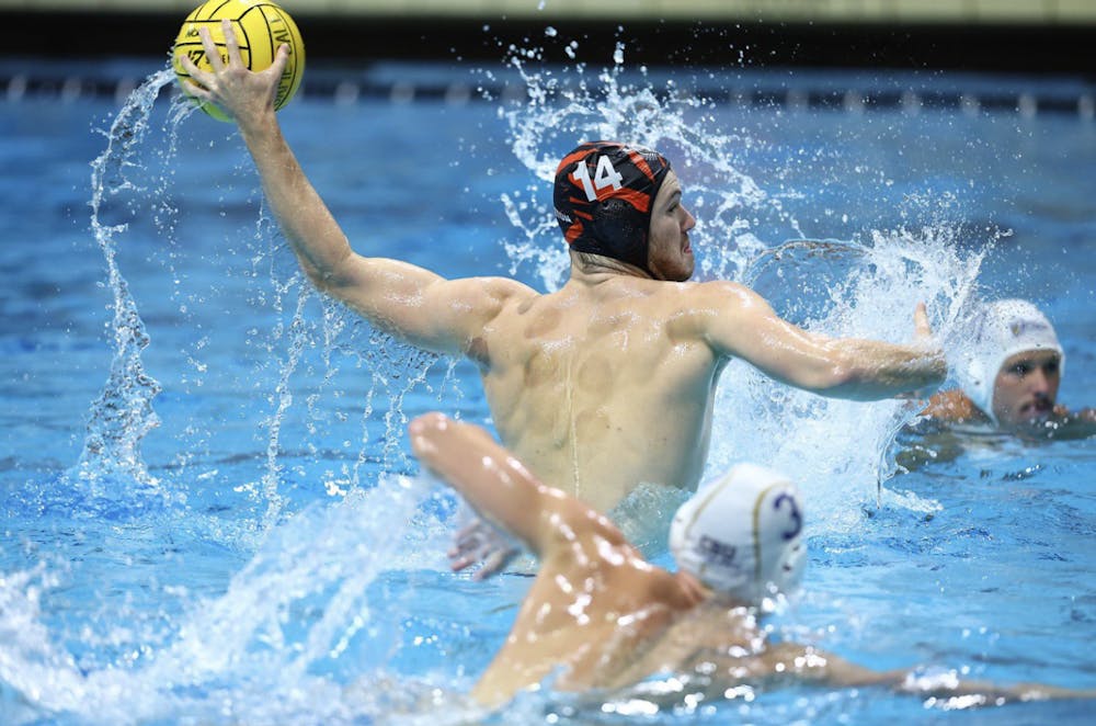 A white man wearing an orange water polo cap with the number 14 throws a water polo ball with two other men in white water polo caps around him. 