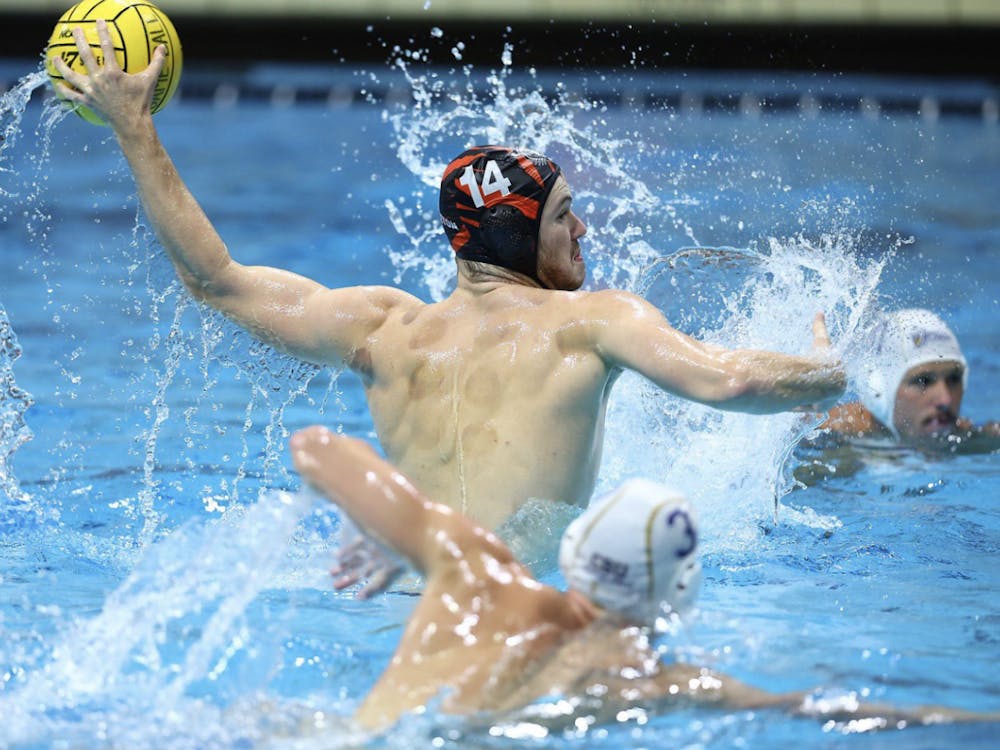 A white man wearing an orange water polo cap with the number 14 throws a water polo ball with two other men in white water polo caps around him. 
