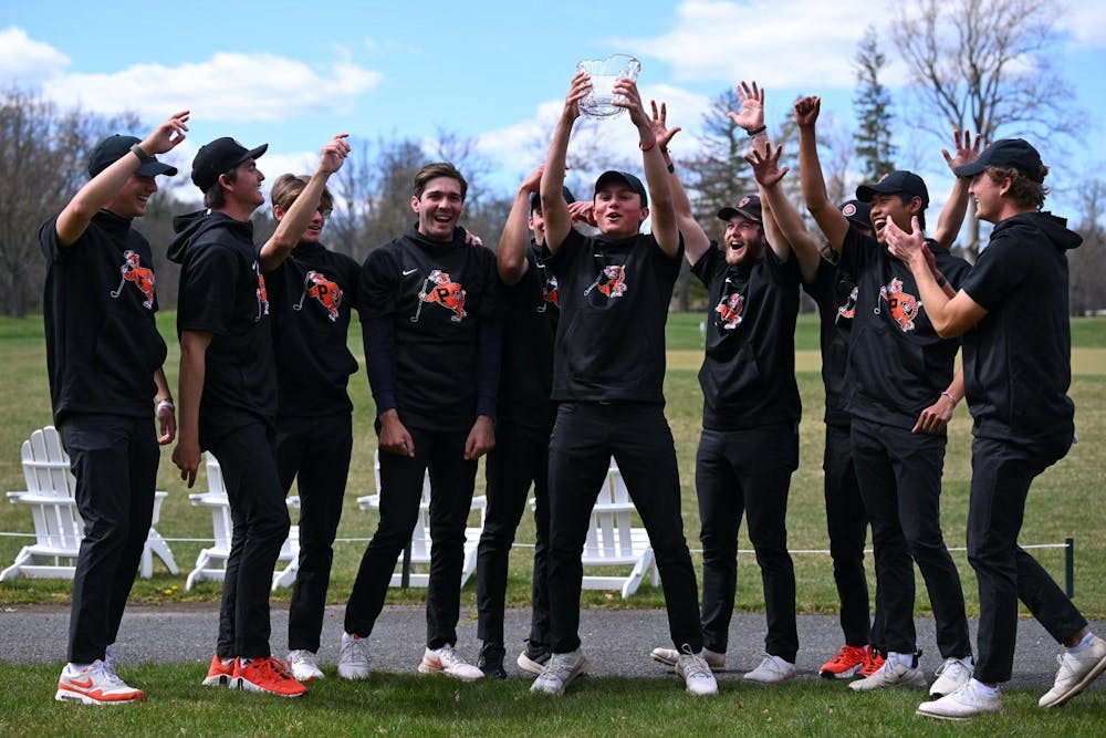 A group of men wearing black attire with a tiger on their shirts celebrate with their hands in the air in front of a grass lawn with one of them holding the trophy. 