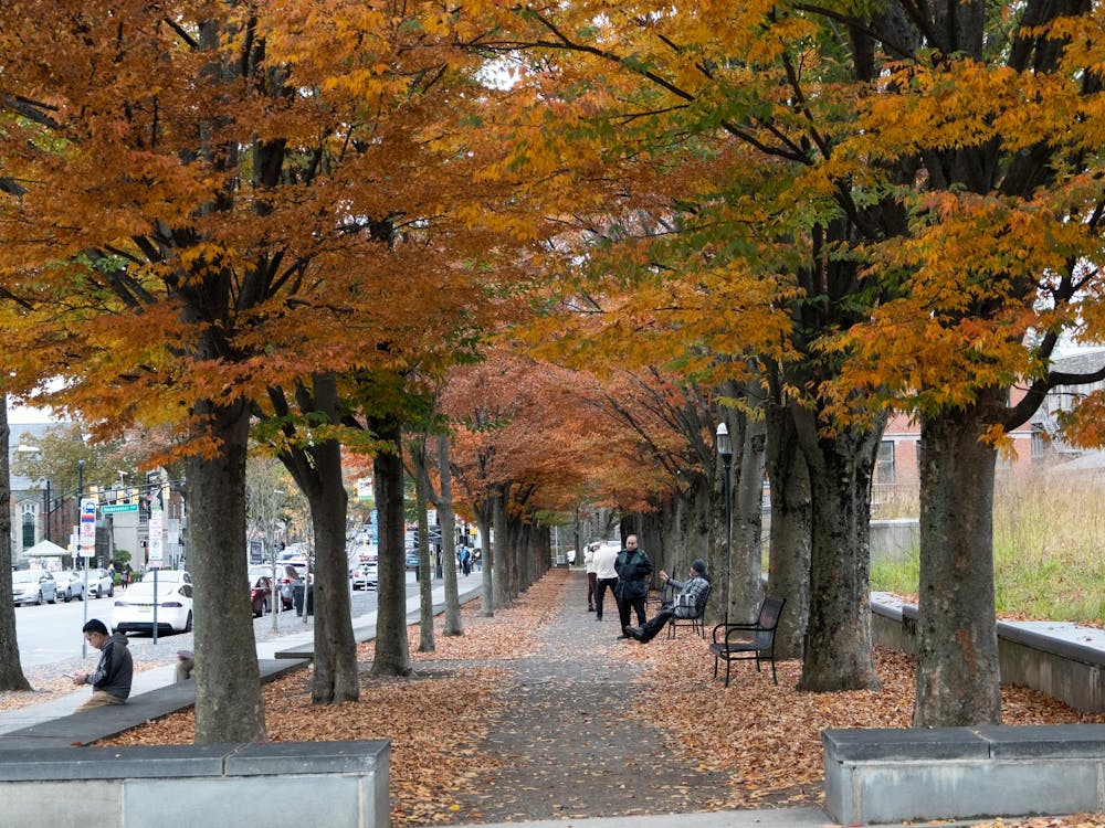 People sit on benches underneath trees that are turning orange and yellow in the Fall. 