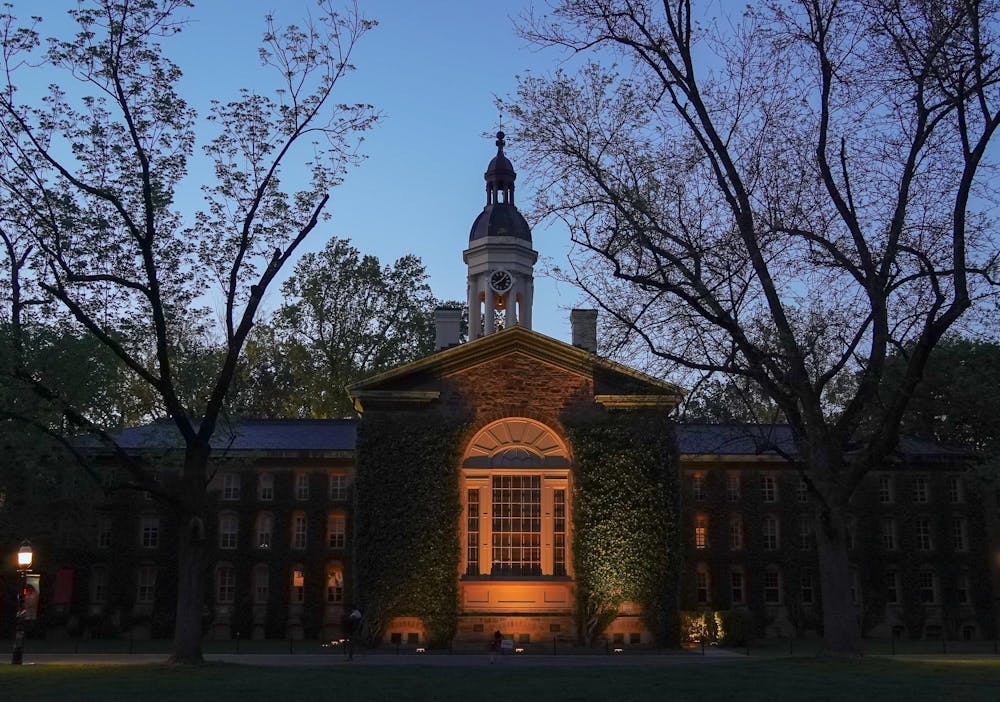 Photo of a building with a clock tower shrouded in ivy, flanked with trees, at dusk.