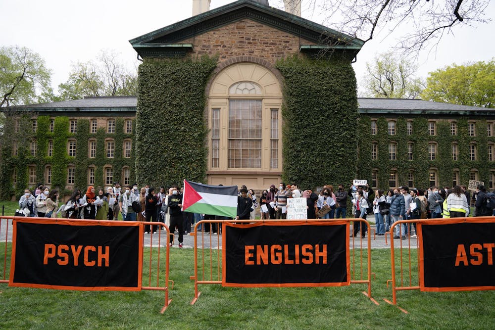 A group of protestors in stand in front of an ivy-covered building, behind three orange and black banners.