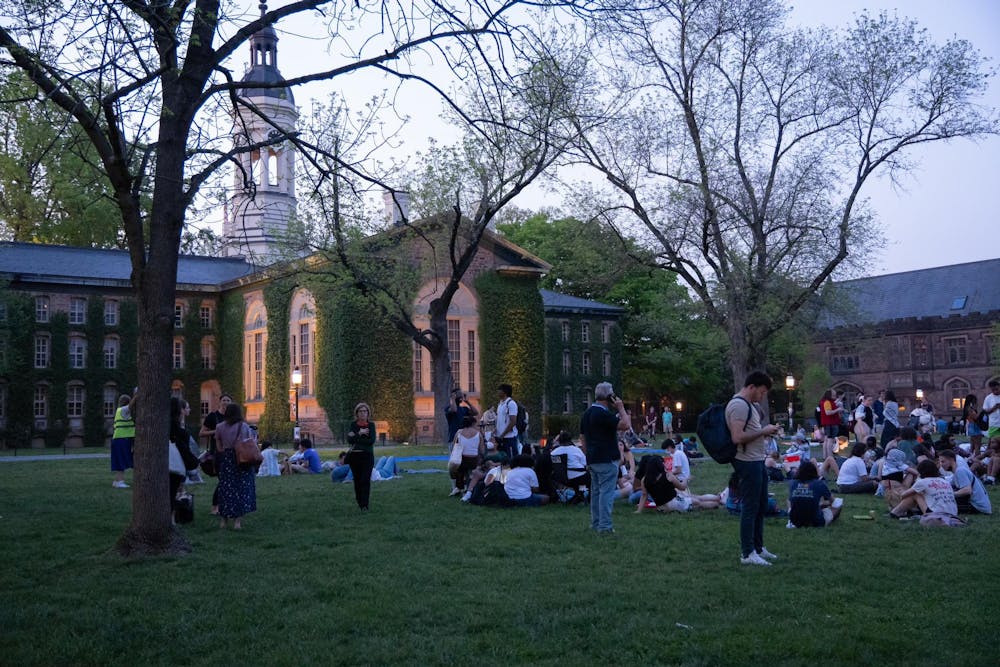 In the background, a brown brick building with green vines growing over it. In the foreground, several individuals sit on green grass.