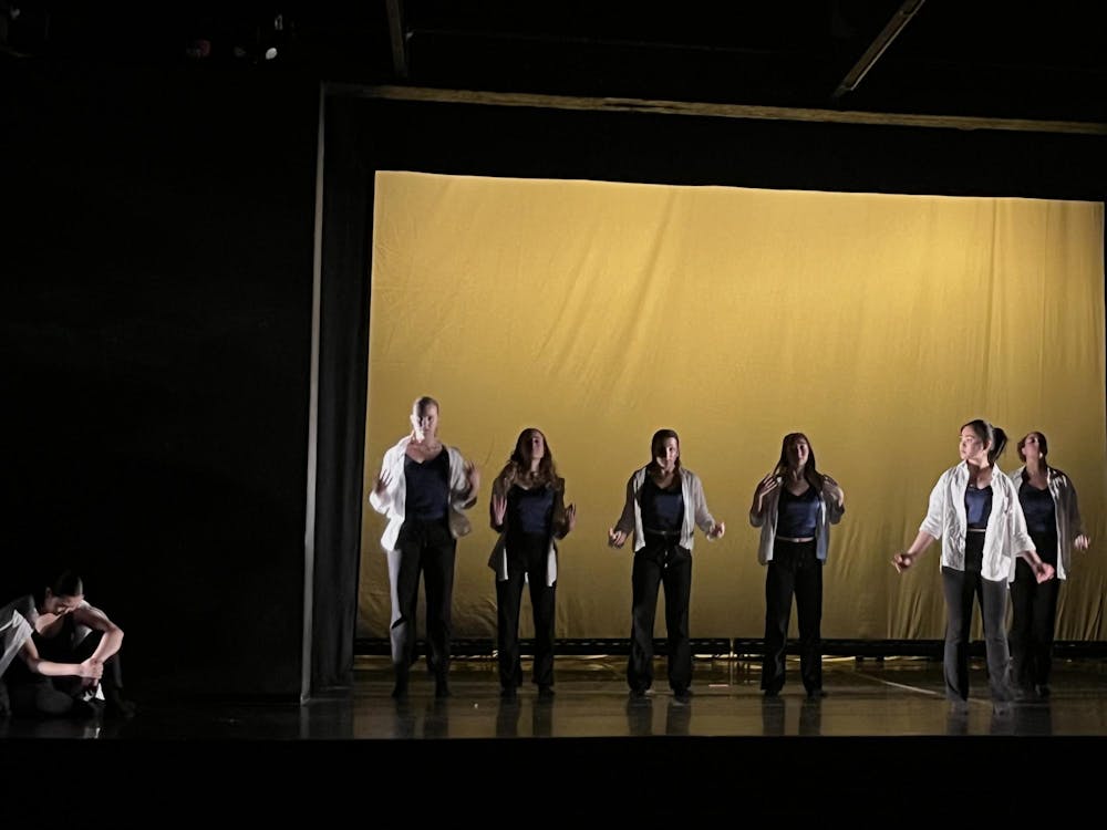 Seven dancers on a dark stage with a yellow backdrop