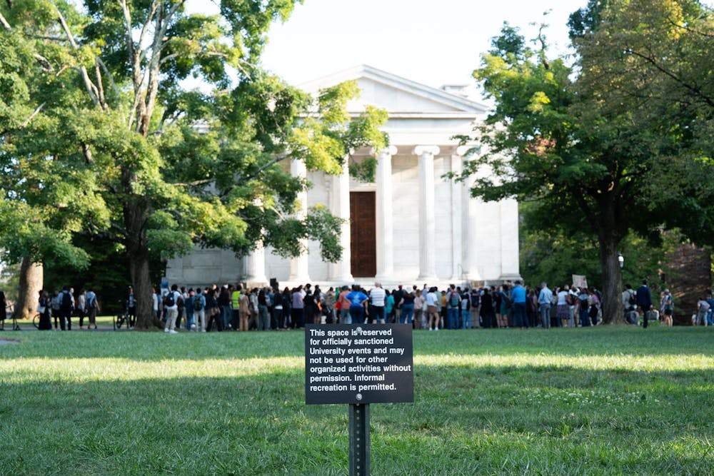 A large white building in the background has two large trees in front of it. A group of people are stood in front of the building. There is a black sign with white writing in the foreground.