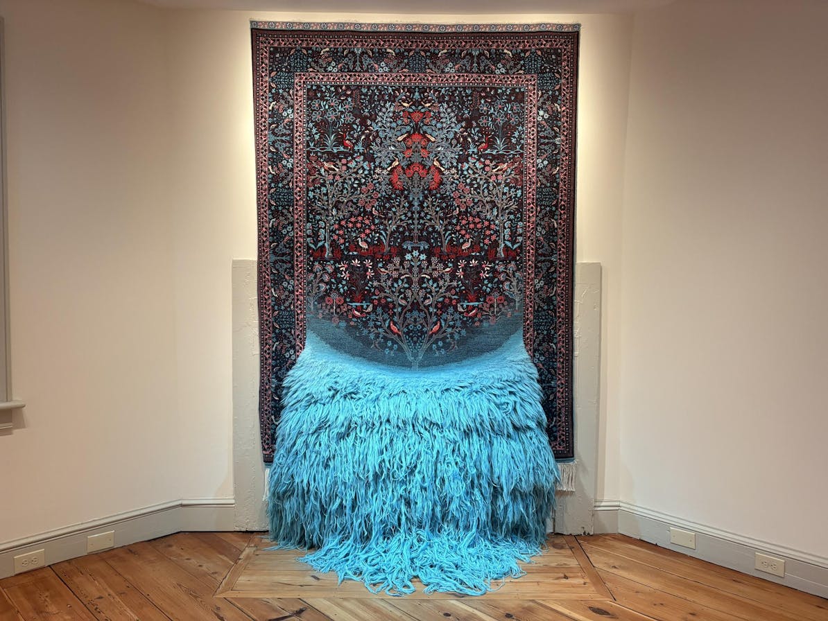 A patterned carpet hangs against a white wall; the top half is a patterned carpet with pink, green, red, and blue embellishments, while the lower portion is an explosion of cyan yarn. 