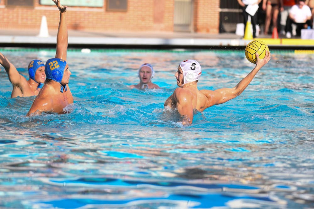 A water polo player with a white cap on in the pool getting ready to shoot the ball with two defenders in blue caps putting their hands to try and block the shot. 