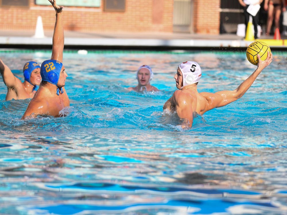 A water polo player with a white cap on in the pool getting ready to shoot the ball with two defenders in blue caps putting their hands to try and block the shot. 
