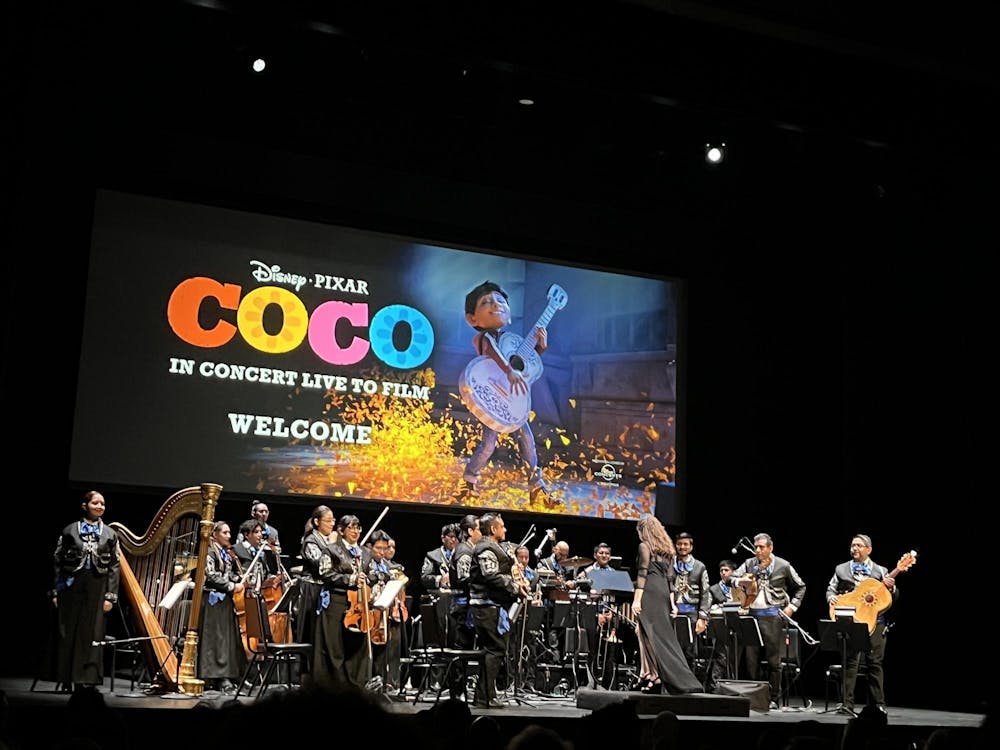 Members of an orchestra stand on the stage of a theater, with the "Coco" movie logo and art projected above them.