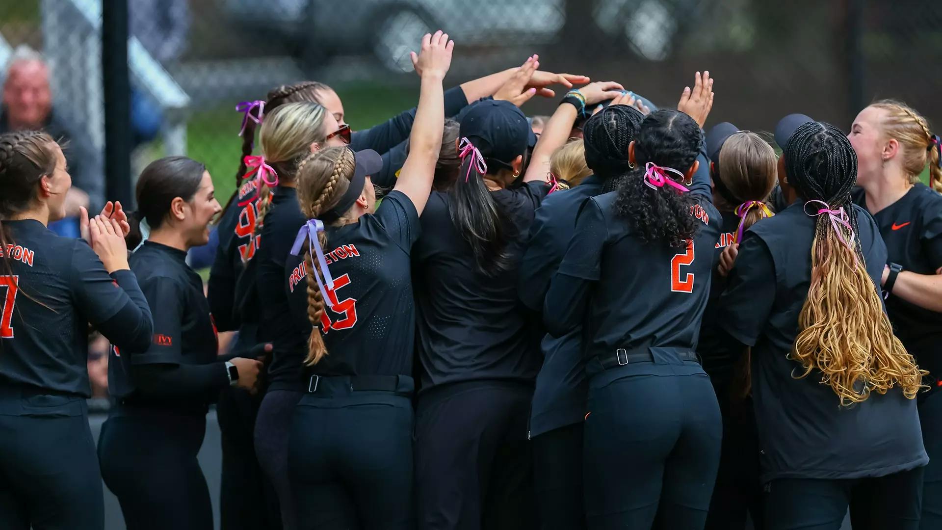 Group of softball players in navy blue and orange uniforms celebrate together around home plate. 