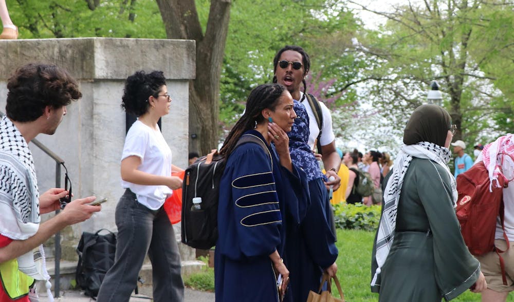 A woman in a blue robe stands facing sideways. Surrouding her are several students walking past.