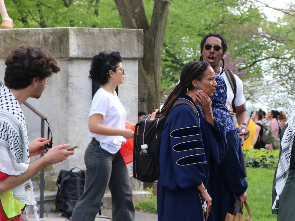 A woman in a blue robe stands facing sideways. Surrouding her are several students walking past.